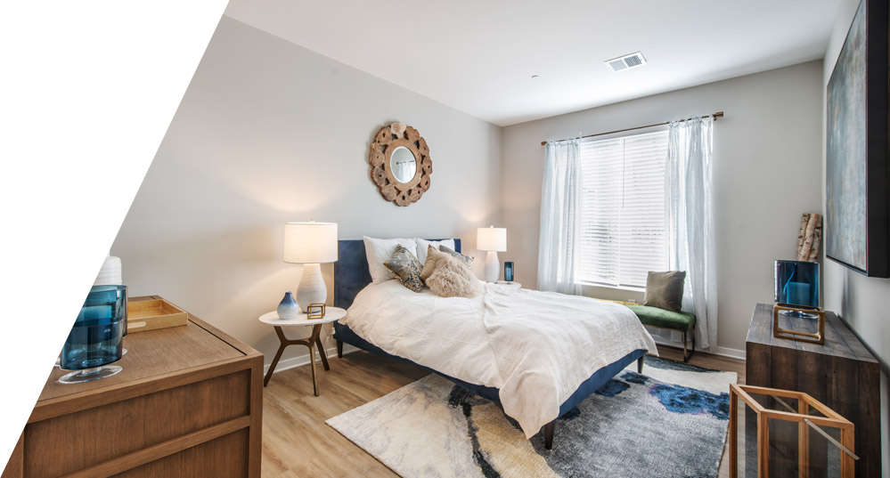 A furnished model apartment bedroom with a window, hardwood-style flooring, and a queen-size bed.